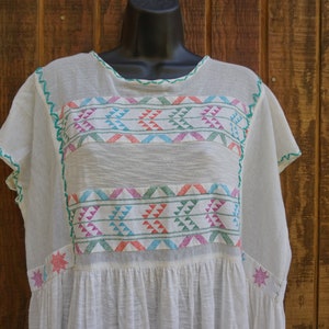 May include: Off-white, short-sleeved tunic with colorful geometric embroidery. The embroidery features a repeating pattern of triangles in shades of pink, blue, and green. The neckline and sleeves are trimmed with green stitching. The tunic is displayed on a black mannequin.
