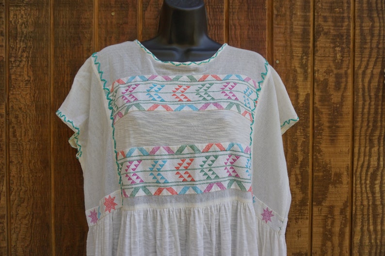 May include: Off-white, short-sleeved tunic with colorful geometric embroidery. The embroidery features a repeating pattern of triangles in shades of pink, blue, and green. The neckline and sleeves are trimmed with green stitching. The tunic is displayed on a black mannequin.