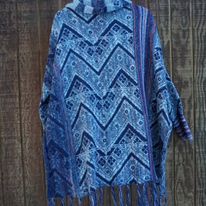 May include: A blue poncho with a geometric pattern and fringe. The poncho features a repeating zigzag design in shades of blue and white. The garment is hanging on a black hanger against a wooden background.