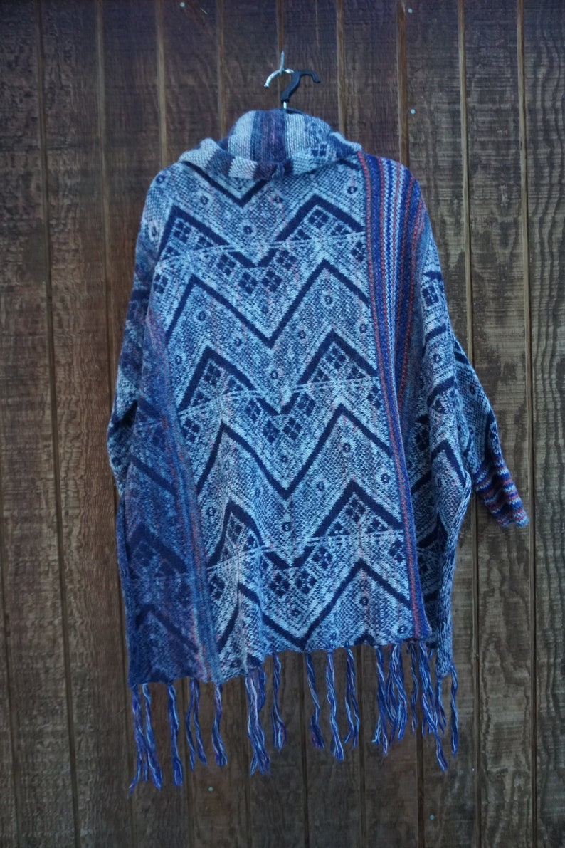 May include: A blue poncho with a geometric pattern and fringe. The poncho features a repeating zigzag design in shades of blue and white. The garment is hanging on a black hanger against a wooden background.