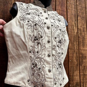 Puede incluir: Chaleco color crema con intrincados bordados y abalorios plateados. El chaleco presenta un cuello alto y una parte delantera abotonada. El dise&ntilde;o incluye patrones florales y arremolinados, creando un aspecto detallado y ornamentado. El chaleco se exhibe en un maniqu&iacute;.