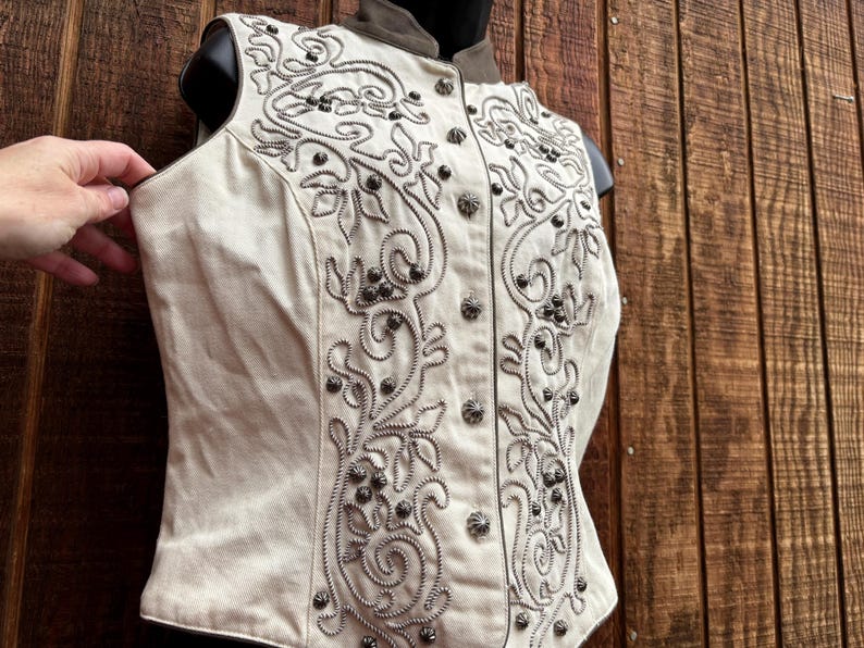 Puede incluir: Chaleco color crema con intrincados bordados y abalorios plateados. El chaleco presenta un cuello alto y una parte delantera abotonada. El dise&ntilde;o incluye patrones florales y arremolinados, creando un aspecto detallado y ornamentado. El chaleco se exhibe en un maniqu&iacute;.