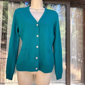 May include: A solid teal long-sleeve cardigan featuring a V-neckline, a front button-down closure with five light-colored buttons, and two patch pockets at the waist. The knit garment has ribbed cuffs and a ribbed hemline for a finished look.