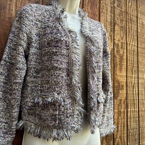 May include: A multi-colored tweed jacket with fringe trim. The jacket features a collarless design, two front pockets, and a fringed hem and edges. The tweed fabric incorporates shades of gray, purple, and cream. The jacket is displayed on a mannequin.