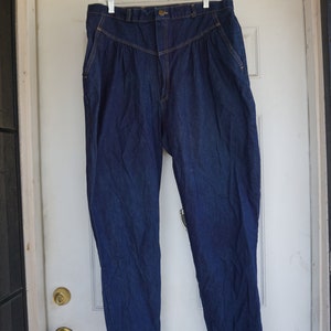 May include: Dark blue denim trousers with a high waist and pleated front. The trousers are hanging on a white hanger against a white door. The trousers have a button closure and pockets.