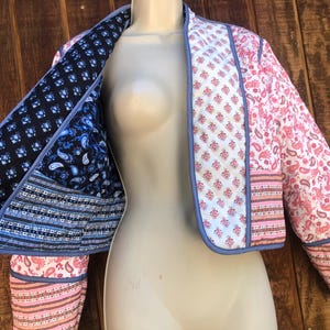 May include: A quilted jacket with a patchwork design. The jacket features a mix of patterns, including floral and paisley prints in pink, white, and blue. The jacket is open, revealing the patterned lining. The edges are trimmed with a light blue binding.