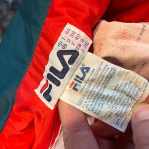 May include: A close-up of a Fila jacket label. The label is white with the Fila logo and text in several languages. The jacket is teal, red, and white. The label includes size information and care instructions.
