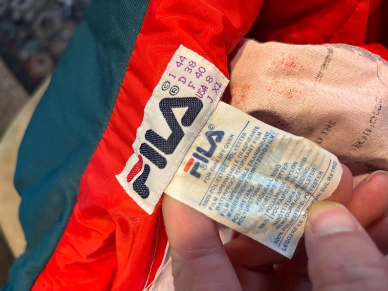 May include: A close-up of a Fila jacket label. The label is white with the Fila logo and text in several languages. The jacket is teal, red, and white. The label includes size information and care instructions.