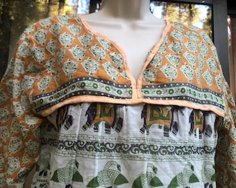 May include: A long-sleeved tunic top with a V-neckline. The top features a pattern of green and white trees on an orange background. The lower portion has a white background with green and black elephant and bird designs. The neckline is trimmed in peach.