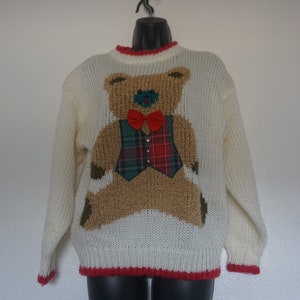 May include: Cream-colored knit sweater featuring a large teddy bear design. The bear has a brown textured body, a plaid vest, and a red bow tie. The sweater has red trim around the collar and cuffs.