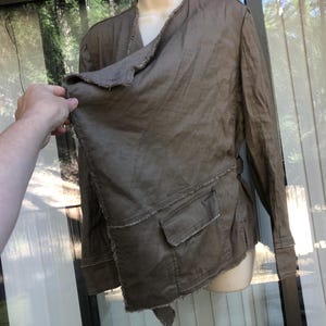 K&ouml;nnte beinhalten: Braune Jacke mit einem einzigartigen asymmetrischen Design. Die Jacke hat lange &Auml;rmel, eine Wickeloptik vorne und zwei Vordertaschen. Die Kanten haben einen ausgefransten, abgenutzten Look. Die Jacke wird auf einer Schaufensterpuppe pr&auml;sentiert.