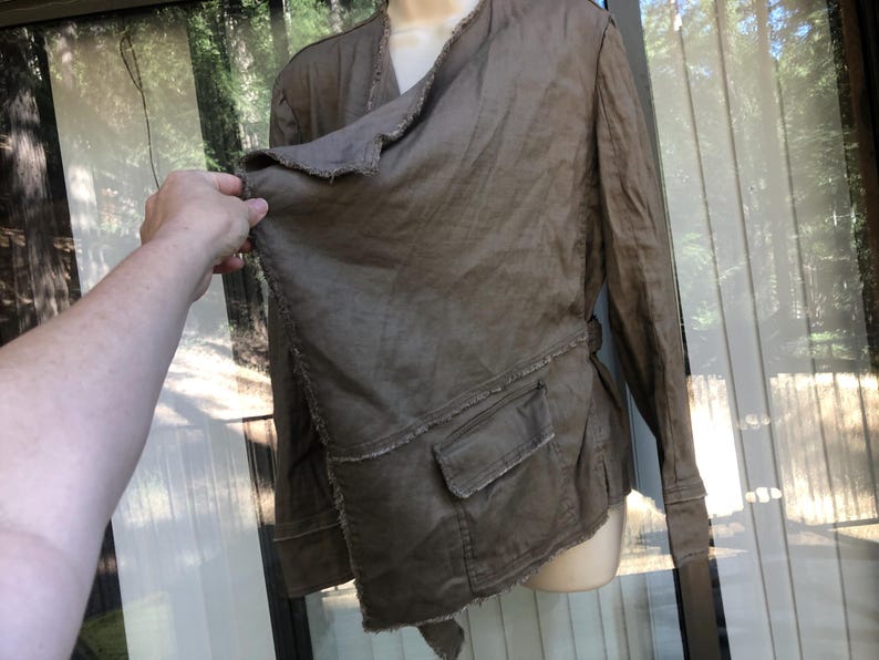K&ouml;nnte beinhalten: Braune Jacke mit einem einzigartigen asymmetrischen Design. Die Jacke hat lange &Auml;rmel, eine Wickeloptik vorne und zwei Vordertaschen. Die Kanten haben einen ausgefransten, abgenutzten Look. Die Jacke wird auf einer Schaufensterpuppe pr&auml;sentiert.