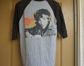 May include: Gray raglan-style t-shirt with black sleeves. The shirt features a black and white image of Bruce Springsteen with the text "BRUCE SPRINGSTEEN AND THE STREET BAND" in orange.