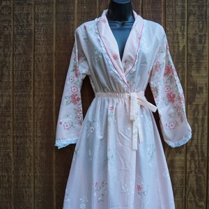 May include: A light pink floral print robe with long sleeves and a matching tie belt. The robe features a delicate floral pattern in shades of pink and green, with white lace trim along the edges. The robe is displayed on a black mannequin.