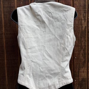 Puede incluir: Un chaleco color crema con cuello marr&oacute;n, exhibido en una percha negra. El chaleco tiene un corte entallado y un dobladillo curvo. El fondo es una pared de madera.