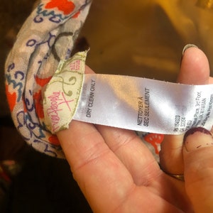 May include: A close-up of a patterned scarf with a tag that reads "Free People" and "Dry Clean Only." The scarf has a floral design with orange and blue accents. The tag also includes the text "NETTOYER A SEC SEULEMENT."