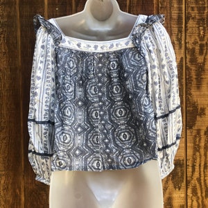 Puede incluir: Una blusa estampada en blanco y azul, con cuello cuadrado y mangas largas. La parte delantera presenta un dise&ntilde;o geom&eacute;trico, mientras que las mangas lucen estampados florales. Volantes y ribetes azul oscuro a&ntilde;aden detalles.