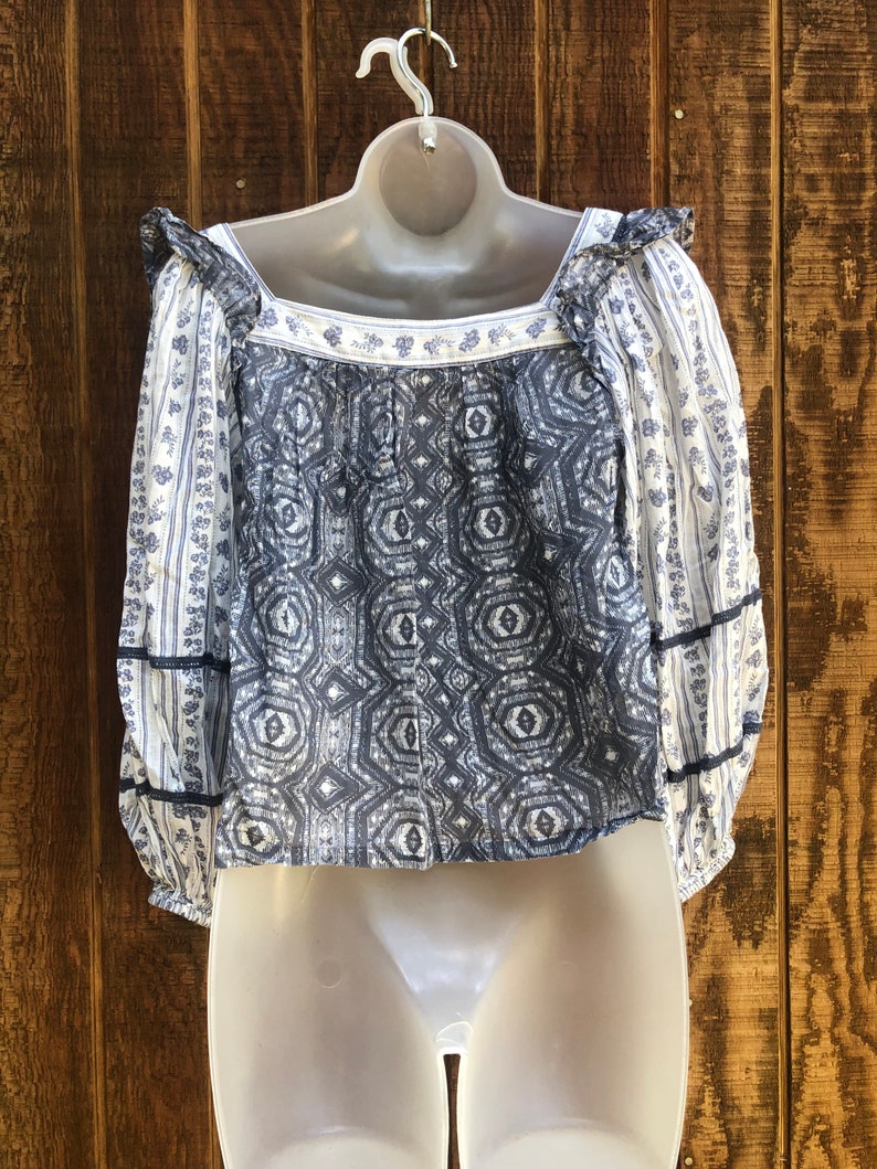 Puede incluir: Una blusa estampada en blanco y azul, con cuello cuadrado y mangas largas. La parte delantera presenta un dise&ntilde;o geom&eacute;trico, mientras que las mangas lucen estampados florales. Volantes y ribetes azul oscuro a&ntilde;aden detalles.