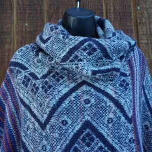 May include: A blue and white patterned poncho with a cowl neck. The poncho features a geometric design in shades of blue, with accents of red and orange. The garment is displayed on a black mannequin head.
