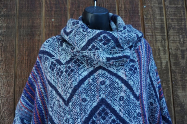 May include: A blue and white patterned poncho with a cowl neck. The poncho features a geometric design in shades of blue, with accents of red and orange. The garment is displayed on a black mannequin head.