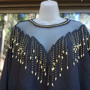 Pode incluir: Camisola preta com decote de malha transparente e detalhes de franjas de contas douradas e pretas.