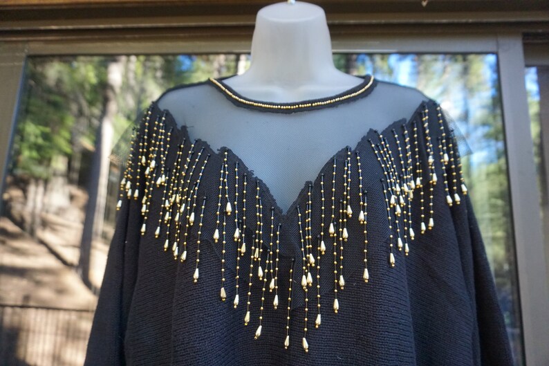 Pode incluir: Camisola preta com decote de malha transparente e detalhes de franjas de contas douradas e pretas.