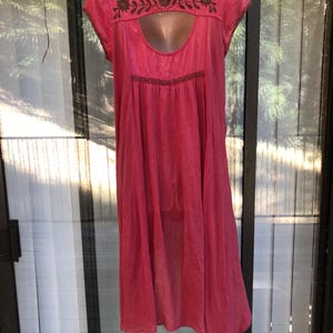 May include: A bright pink dress with short sleeves and a keyhole neckline. The dress features brown embroidered floral detailing around the neckline and bodice. The dress is made of a lightweight fabric and has a flowing, loose fit.