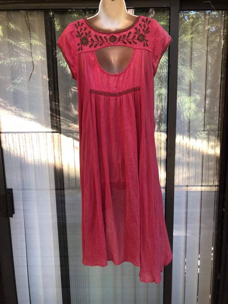 May include: A bright pink dress with short sleeves and a keyhole neckline. The dress features brown embroidered floral detailing around the neckline and bodice. The dress is made of a lightweight fabric and has a flowing, loose fit.