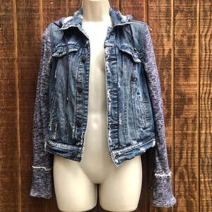 May include: A blue denim jacket with a distressed look, featuring a hood and knit sleeves. The jacket has a button-up front and is displayed on a mannequin against a wooden background. The sleeves are a heathered blue color, and the jacket has a classic, casual style.