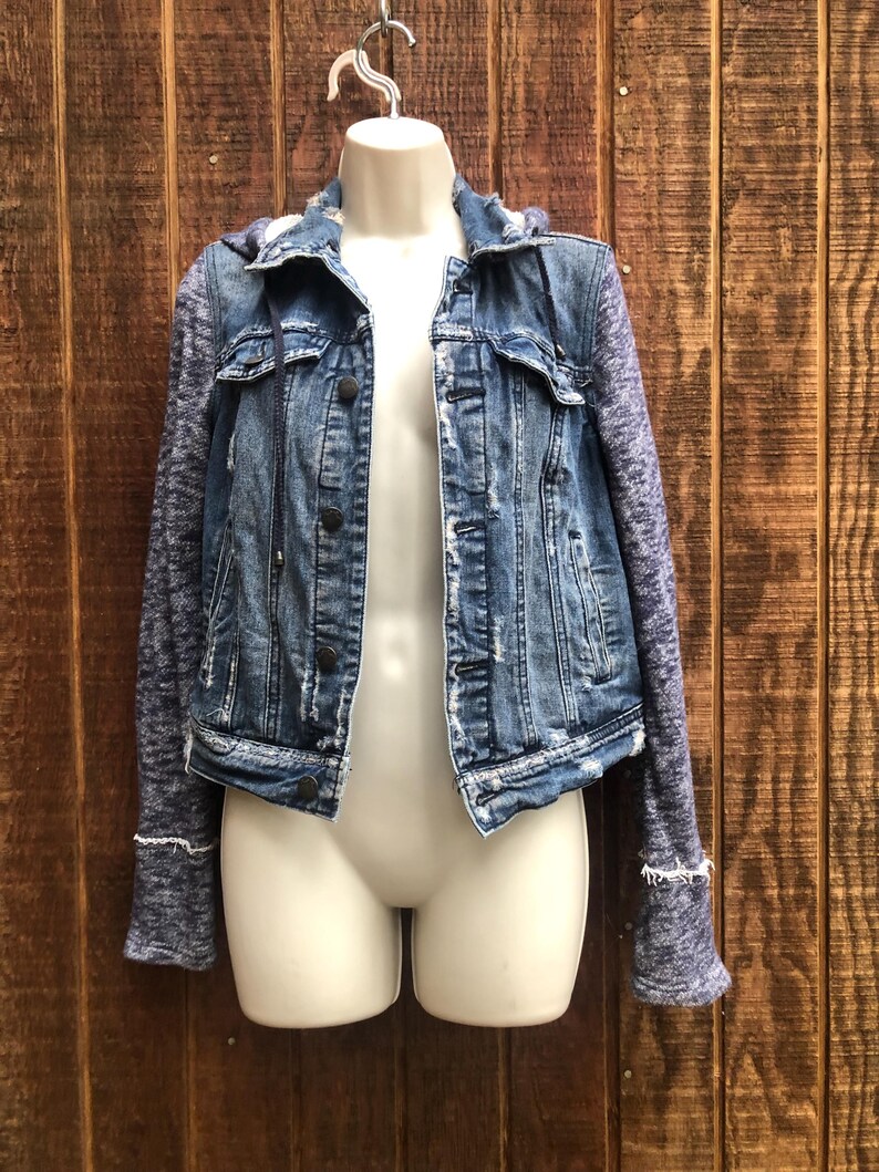 May include: A blue denim jacket with a distressed look, featuring a hood and knit sleeves. The jacket has a button-up front and is displayed on a mannequin against a wooden background. The sleeves are a heathered blue color, and the jacket has a classic, casual style.