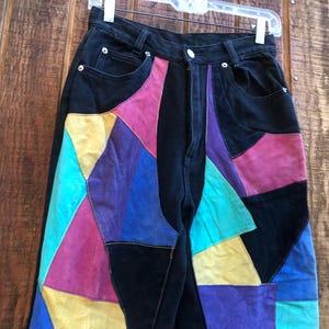 May include: Black denim jeans with colorful patchwork panels in shades of teal, purple, pink, and yellow. The jeans have a classic five-pocket design and a zipper fly.