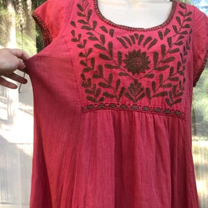 May include: A vibrant pink dress with short sleeves and a scoop neckline. The dress features intricate brown embroidered floral designs across the chest and neckline. The fabric has a textured appearance, and the dress appears to be lightweight and flowing.