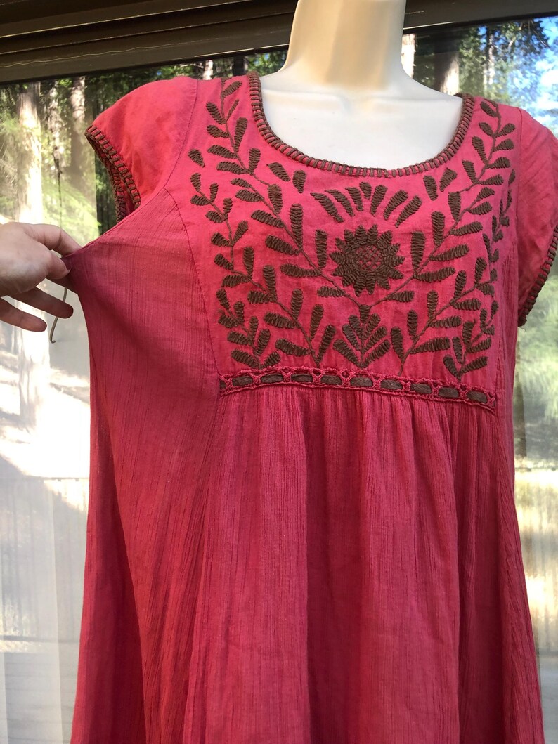May include: A vibrant pink dress with short sleeves and a scoop neckline. The dress features intricate brown embroidered floral designs across the chest and neckline. The fabric has a textured appearance, and the dress appears to be lightweight and flowing.