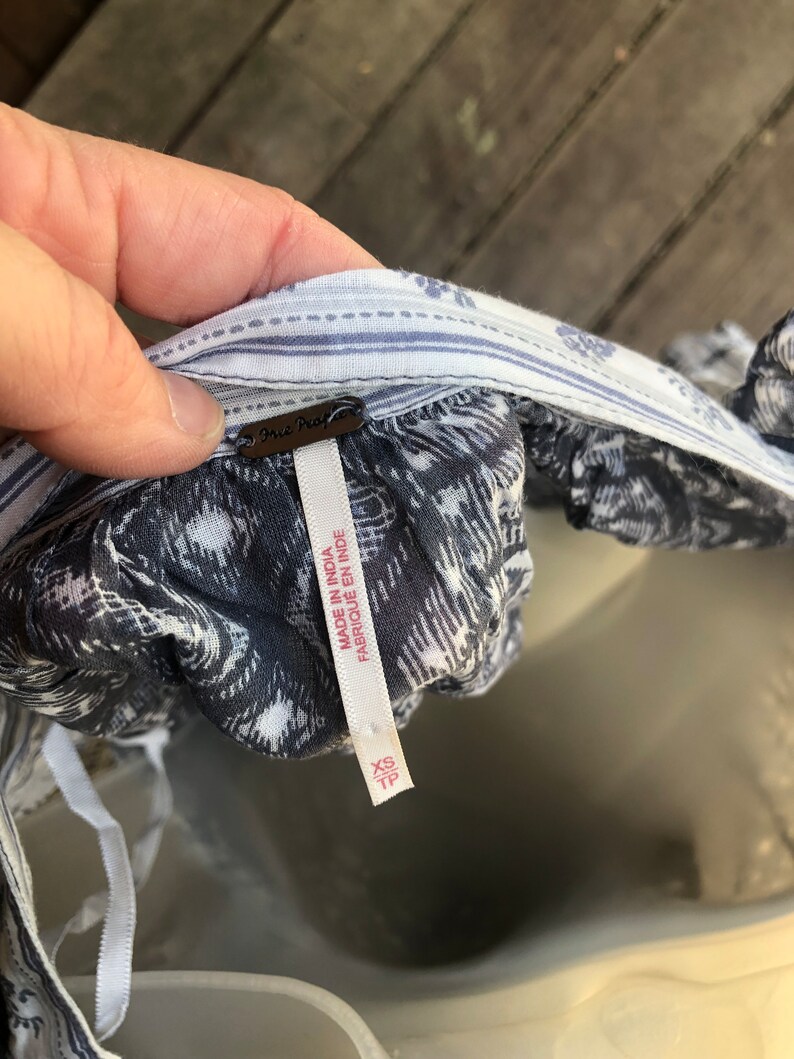 Puede incluir: Primer plano de una blusa estampada azul y blanca con una etiqueta que dice "Free People". La etiqueta tambi&eacute;n dice "Made in India" y "XS/TP". La blusa tiene un escote a rayas.