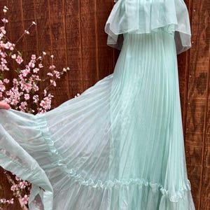 May include: A floor-length mint green dress featuring a pleated skirt, a ruffled bodice overlay, and thin spaghetti straps. The sheer fabric creates a layered effect, with a ruffled hemline adding volume to the flowing silhouette of the garment.