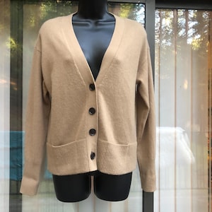 May include: A tan cardigan sweater with a V-neck, long sleeves, and two front pockets. The sweater has four black buttons down the front. The garment is displayed on a black mannequin.