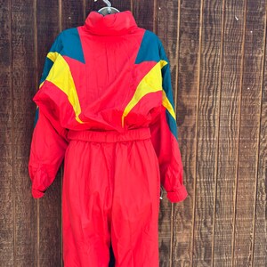 May include: A vintage red ski suit with teal and yellow detailing. The one-piece suit has a high collar and a cinched waist. The jacket features a colour-blocked design with yellow and teal geometric shapes. The suit is hanging against a wooden background.