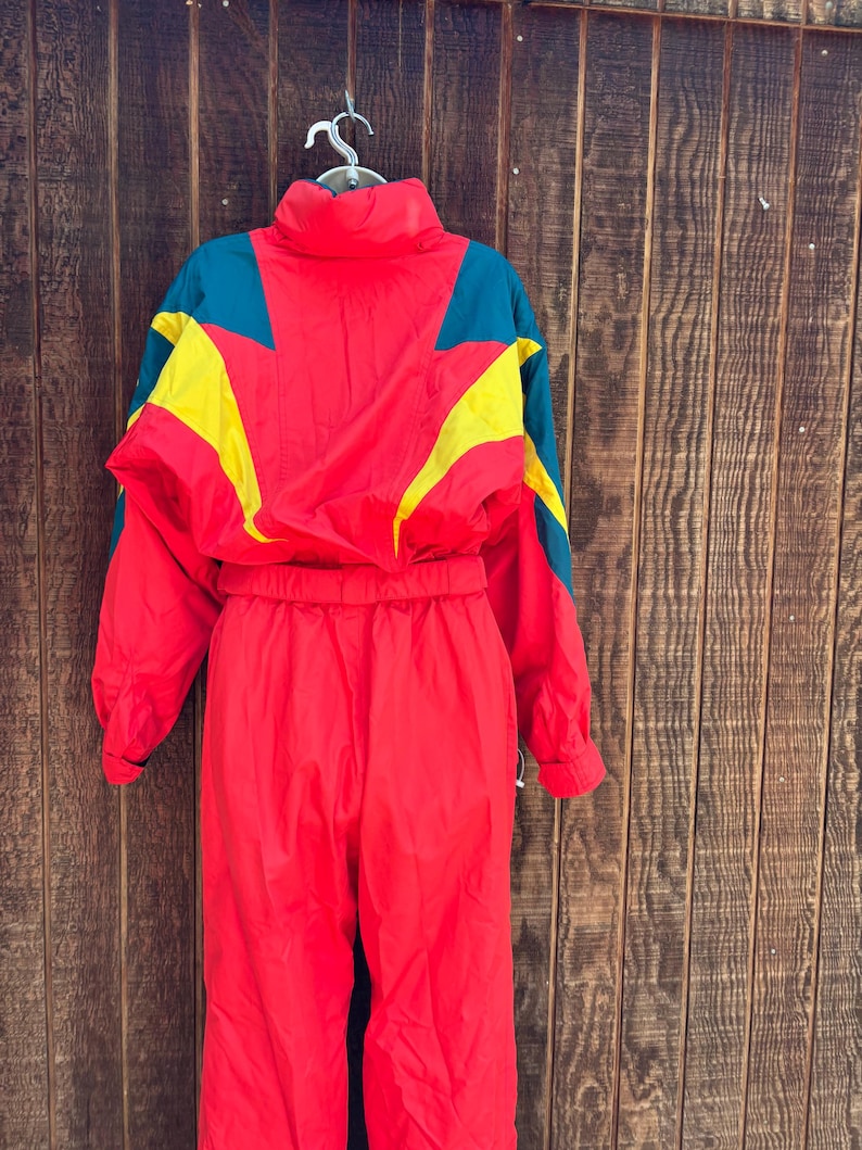 May include: A vintage red ski suit with teal and yellow detailing. The one-piece suit has a high collar and a cinched waist. The jacket features a colour-blocked design with yellow and teal geometric shapes. The suit is hanging against a wooden background.