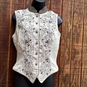 Puede incluir: Chaleco color crema con intrincados patrones florales bordados y detalles de tachuelas plateadas. El chaleco presenta un cuello alto y una parte delantera abotonada. El chaleco se exhibe sobre un maniqu&iacute; negro sobre un fondo de madera.