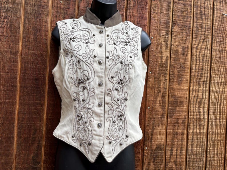 Puede incluir: Chaleco color crema con intrincados patrones florales bordados y detalles de tachuelas plateadas. El chaleco presenta un cuello alto y una parte delantera abotonada. El chaleco se exhibe sobre un maniqu&iacute; negro sobre un fondo de madera.