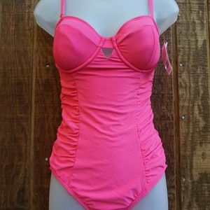 May include: A bright pink one-piece swimsuit displayed on a mannequin. The swimsuit features a sweetheart neckline, adjustable straps, and ruched detailing along the sides. A small tag hangs from the strap. The background is a wooden surface.