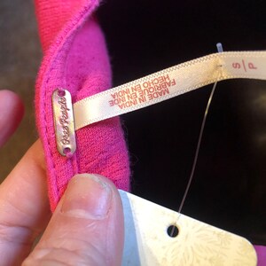 May include: Close-up of a bright pink garment with a small metal tag that reads "Free People". A white tag with the text "MADE IN INDIA" and "S/P" is attached. The garment is made in India.
