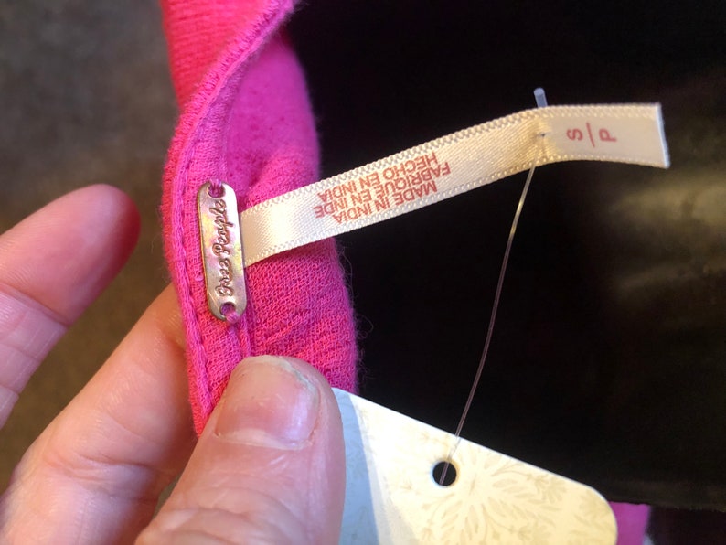 May include: Close-up of a bright pink garment with a small metal tag that reads "Free People". A white tag with the text "MADE IN INDIA" and "S/P" is attached. The garment is made in India.