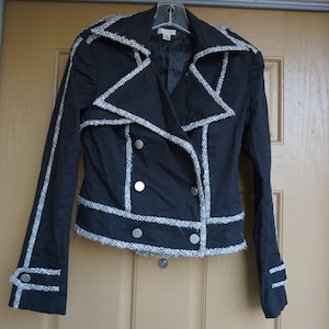 May include: Black jacket with white trim and silver buttons. The jacket has a double-breasted design with a notched collar and long sleeves. The trim is a textured, woven fabric. The jacket is hanging on a hanger against a light brown door.