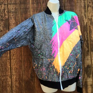 May include: A gray, black, pink, green, and yellow paint splatter patterned jacket with a white zipper. The jacket has a black collar and black cuffs.