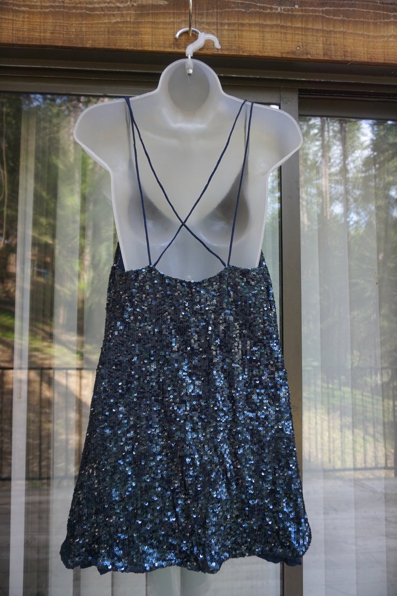 K&ouml;nnte beinhalten: Ein marineblaues Paillettenkleid mit Spaghettitr&auml;gern und tiefem R&uuml;ckenausschnitt h&auml;ngt an einer wei&szlig;en Schaufensterpuppe. Das Kleid ist mit kleinen, reflektierenden Pailletten bedeckt, die einen schimmernden Effekt erzeugen.