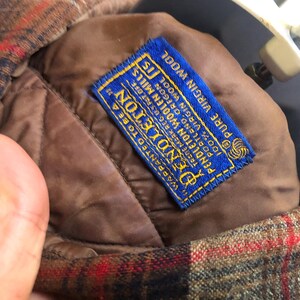 May include: Close-up of a Pendleton wool jacket interior. The jacket features a brown quilted lining and a blue label with gold lettering. The label reads "Pendleton Woolen Mills" and "100% Virgin Wool". The exterior has a red, brown, and black plaid pattern.
