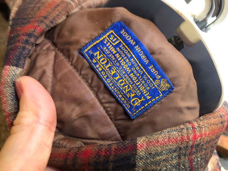 May include: Close-up of a Pendleton wool jacket interior. The jacket features a brown quilted lining and a blue label with gold lettering. The label reads "Pendleton Woolen Mills" and "100% Virgin Wool". The exterior has a red, brown, and black plaid pattern.