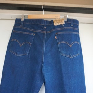 May include: Dark blue denim jeans with orange stitching and classic back pocket design. The jeans are hanging on a wooden hanger. A small tag is visible at the back of the waistband.