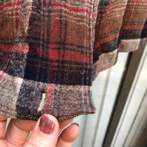 May include: Close-up of a brown, red, and navy blue plaid fabric. The fabric has a textured appearance with visible woven threads. The hem of the garment is visible, with a small opening. The overall color palette is warm and rustic.